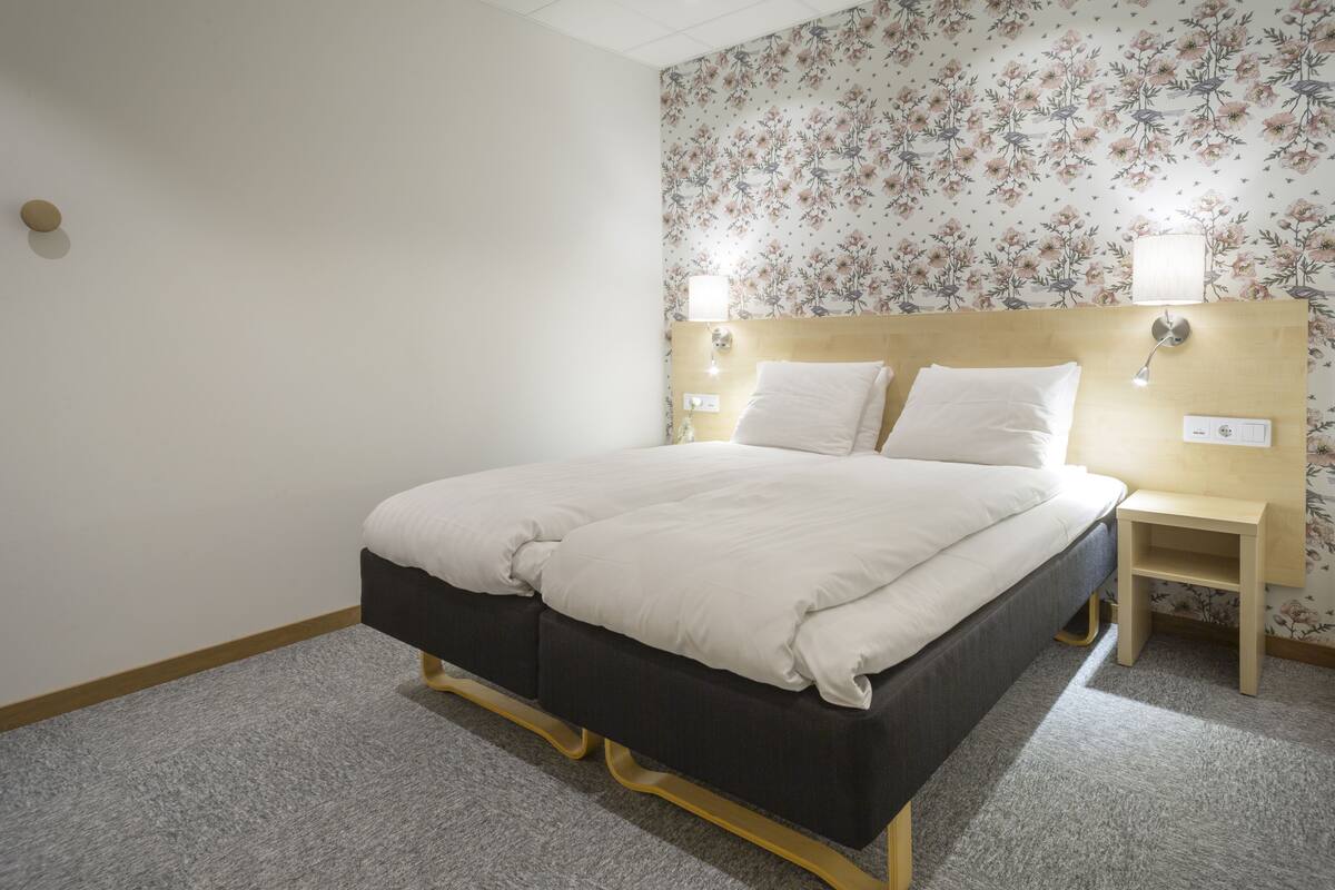 A neatly made bed with a wooden headboard, two bedside tables with lamps, and a floral wallpaper pattern on the wall.