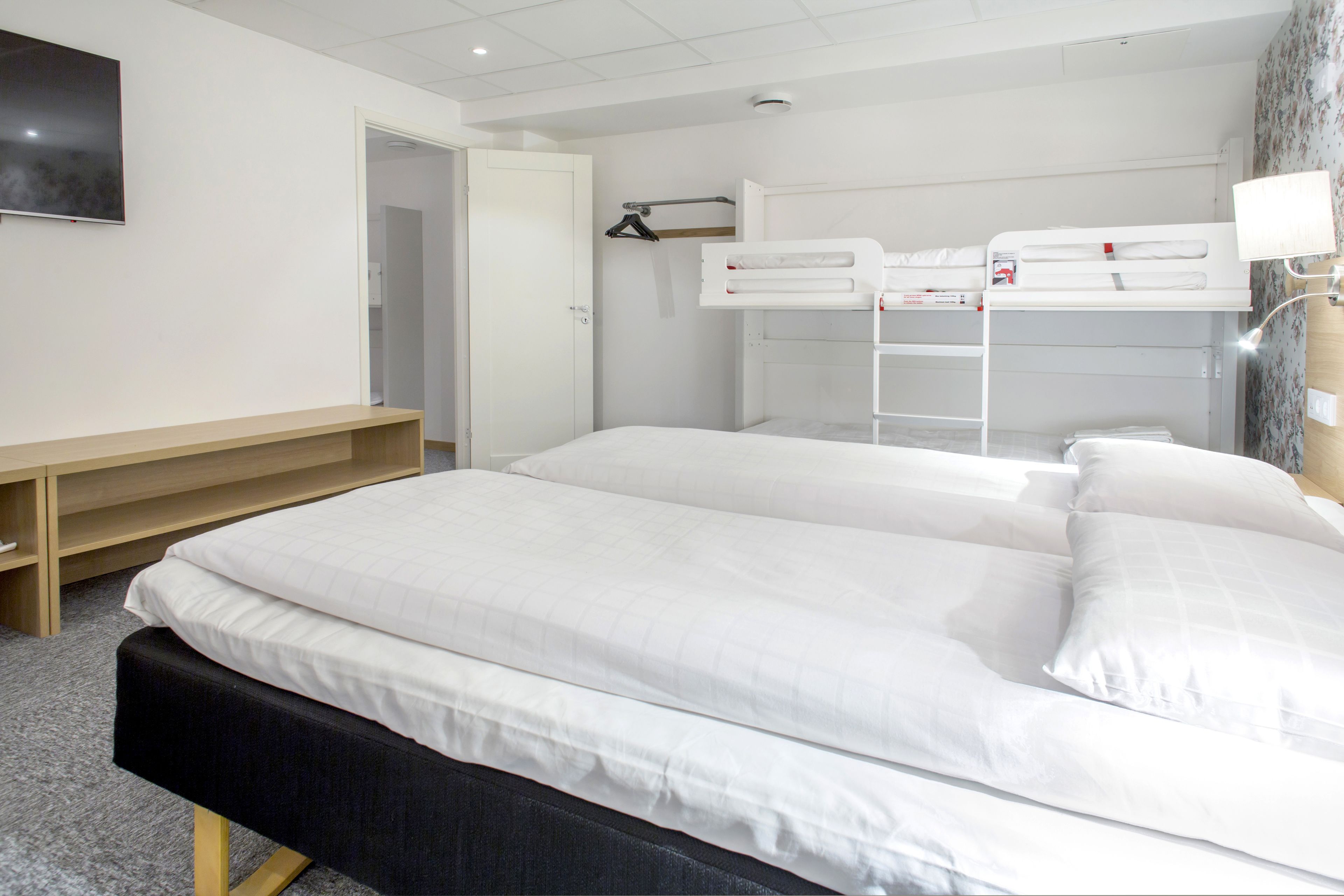 A hotel room with a large bed, a bedside table, a desk, and a bunk bed.