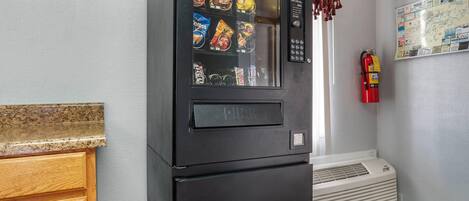 Et køkken med en sort automtisk salgsautomat fyldt med snacks, en brandslukker og et kort på væggen.