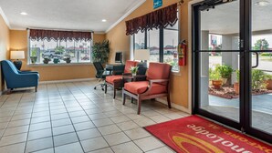 A hotel lobby with a reception desk, two red armchairs, a blue chair, a large window with a view of the parking lot, and a red carpet with the hotel name. - Econo Lodge Oxford near Choccolocco Park (Oxford)
