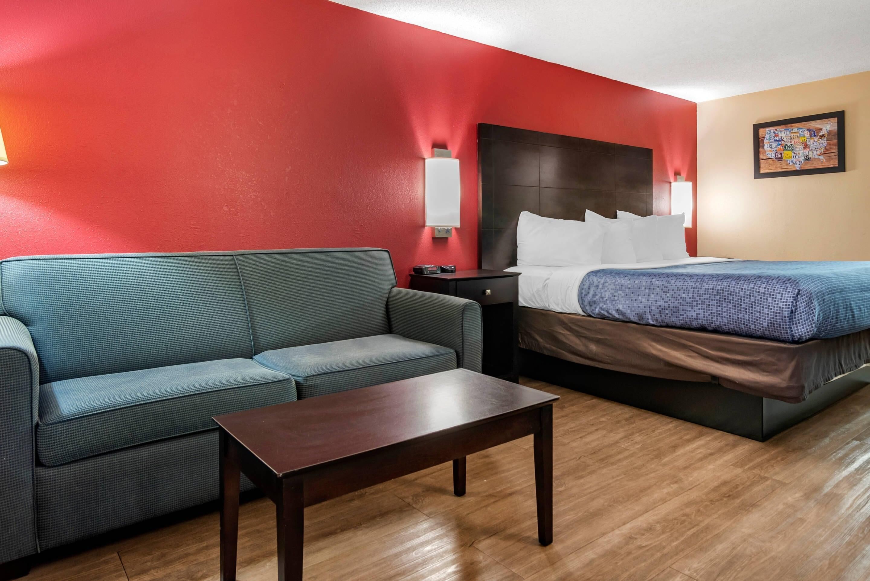 Ein Hotelzimmer mit roter Wand, einem Bett mit weißer Bettwäsche, einem blauen Sofa, einem Holztisch und einem Nachttisch mit einer Lampe.