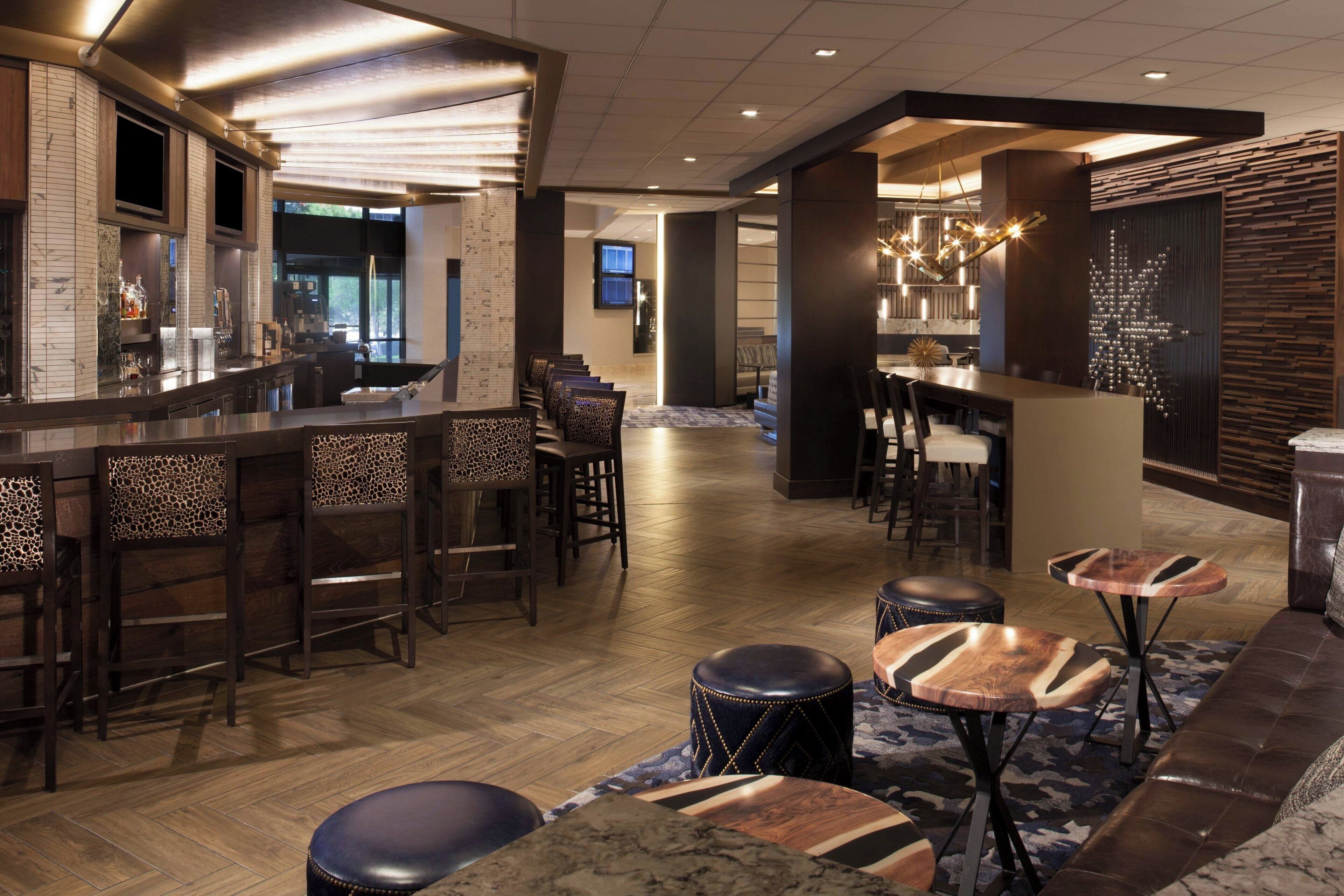 Eine Bar mit hölzernen Barhocker, eine Sitzecke mit einer Couch und runden Tischen sowie ein gemustertes Teppich.