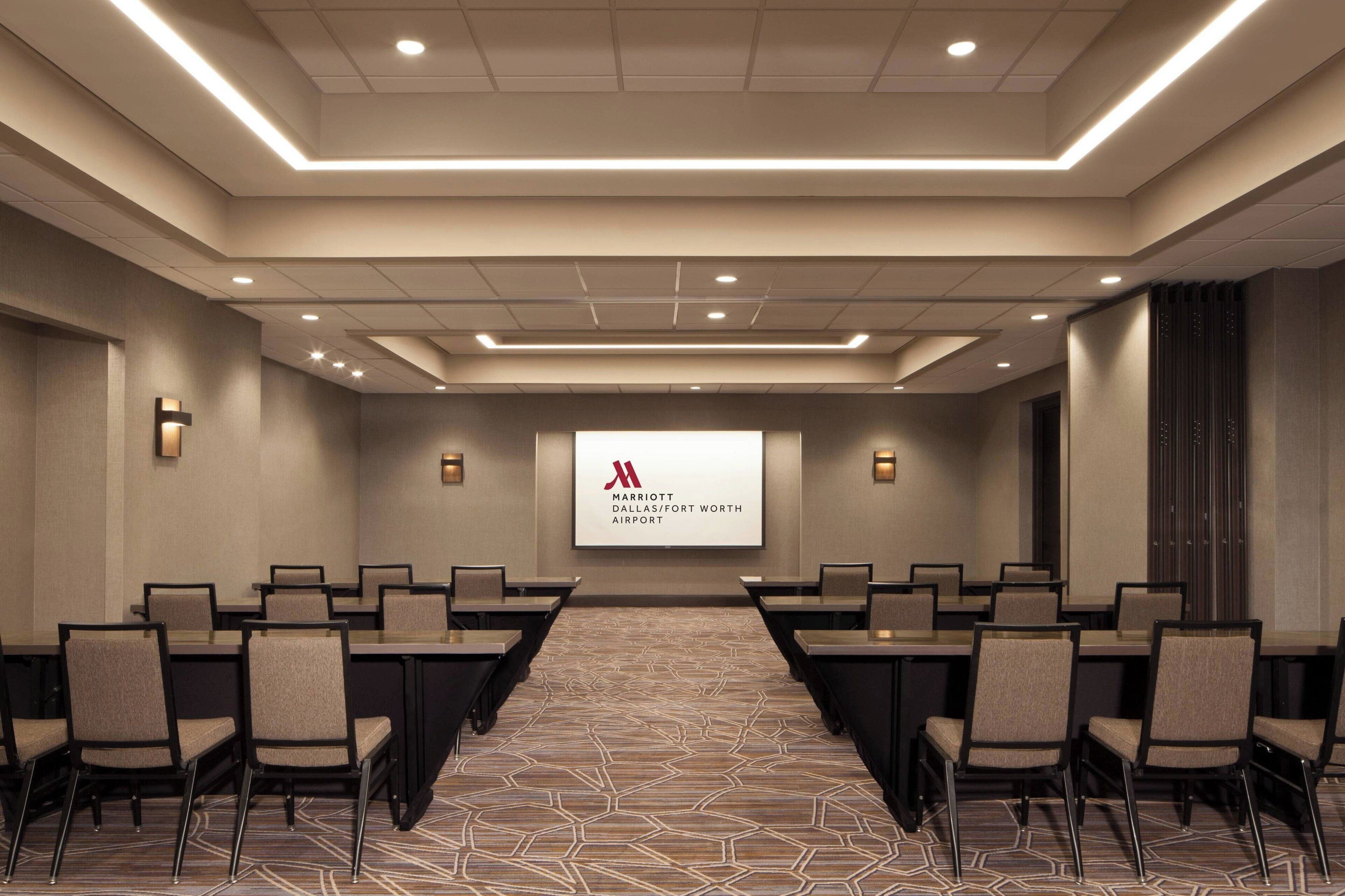 Ein Konferenzraum mit einer großen Leinwand, die das Marriott-Logo zeigt, mehreren Sitzreihen und einem Teppichboden.