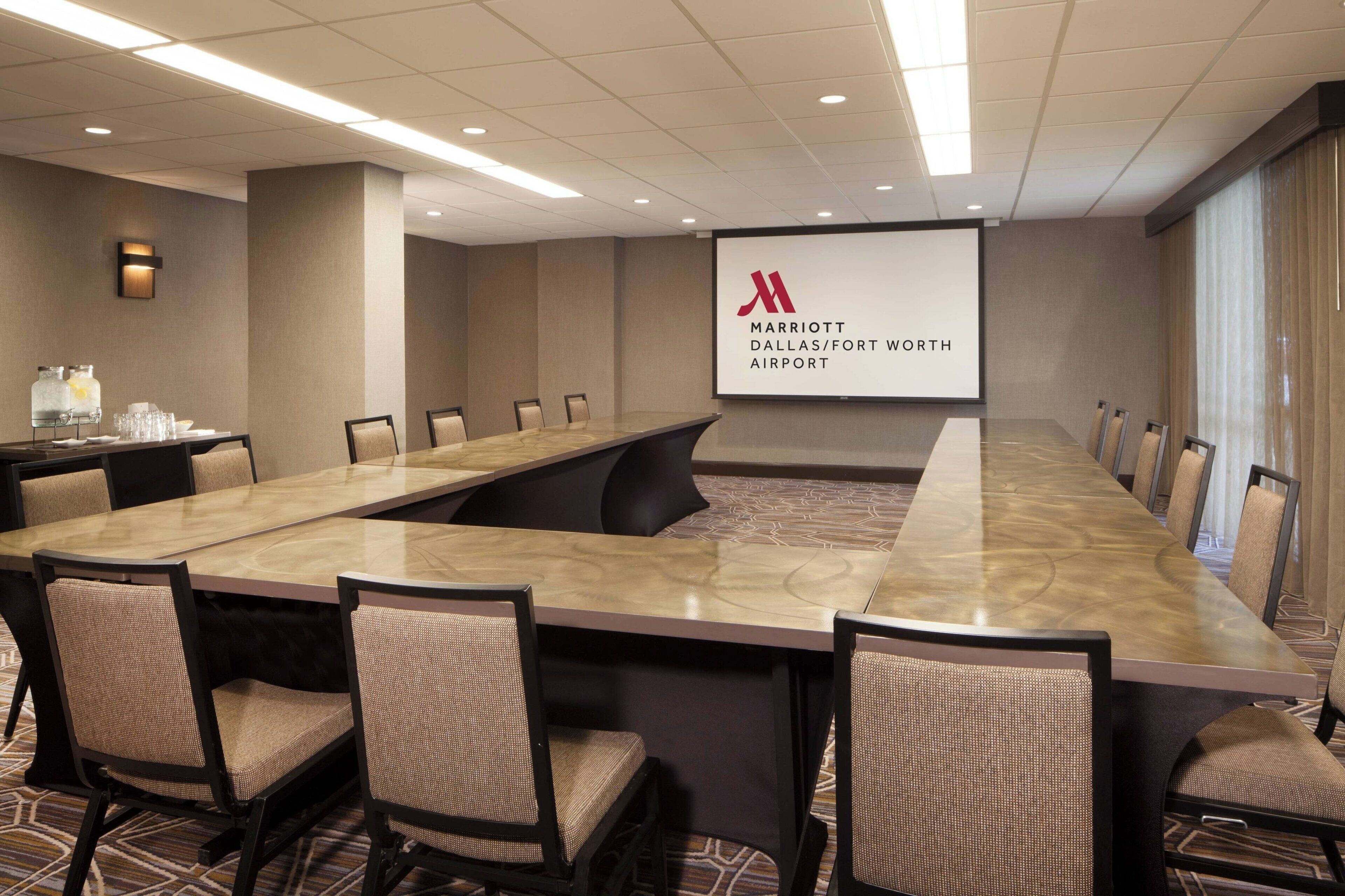 Un salón de conferencias con una larga mesa, sillas, una pantalla de proyector que muestra el logotipo de Marriott Dallas/Fort Worth Airport y un dispensador de agua.