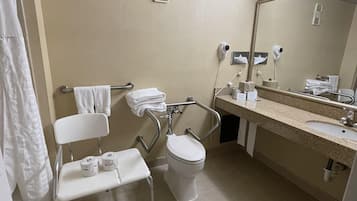 A standard hotel bathroom with a toilet, sink, and hand dryer. There is a wheelchair-accessible bench and a towel rack.