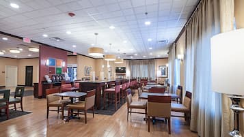 Ampla área de jantar de hotel com várias mesas e cadeiras, um balcão com caixa registradora e vista para a cidade através de grandes janelas.