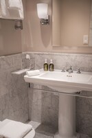Un baño con lavamanos blanco, paredes de mármol, un perchero para toallas, un dispensador de jabón de vidrio y una caja de pañuelos.