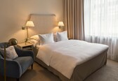A hotel room with a large bed, two bedside lamps, a nightstand, a blue armchair, and a window with sheer curtains.