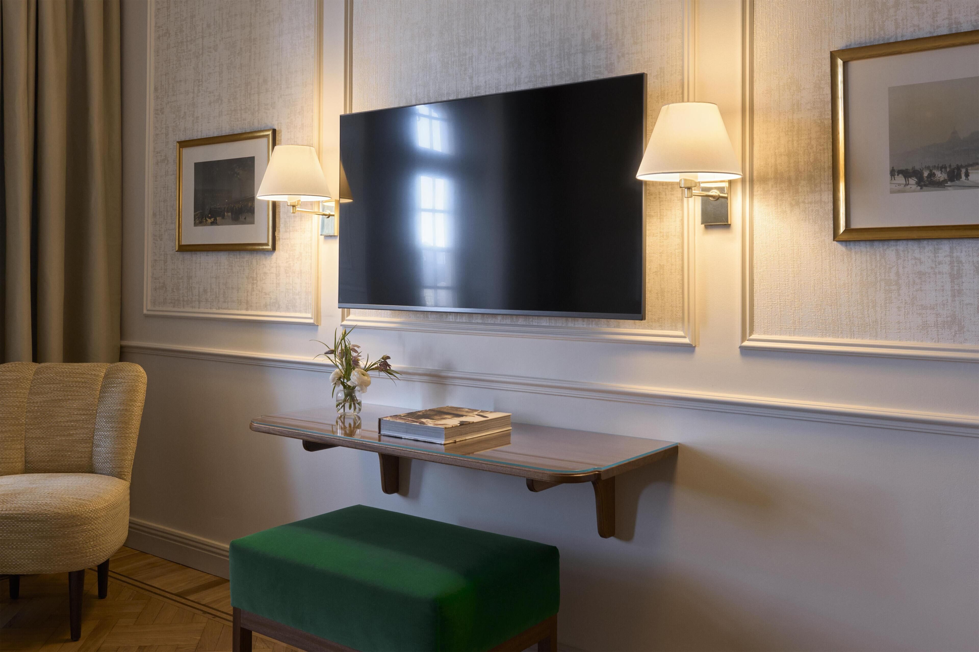 A room with a flat-screen TV mounted on the wall, a small wooden shelf with a book and a vase, a green ottoman, and a chair.