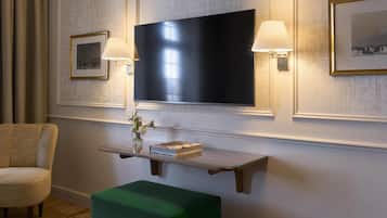 A room with a flat-screen TV mounted on the wall, a small wooden shelf with a book and a vase, a green ottoman, and a chair.