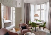 A room with a bed, a pink armchair, a green armchair, a round table with chairs, and a window with curtains.