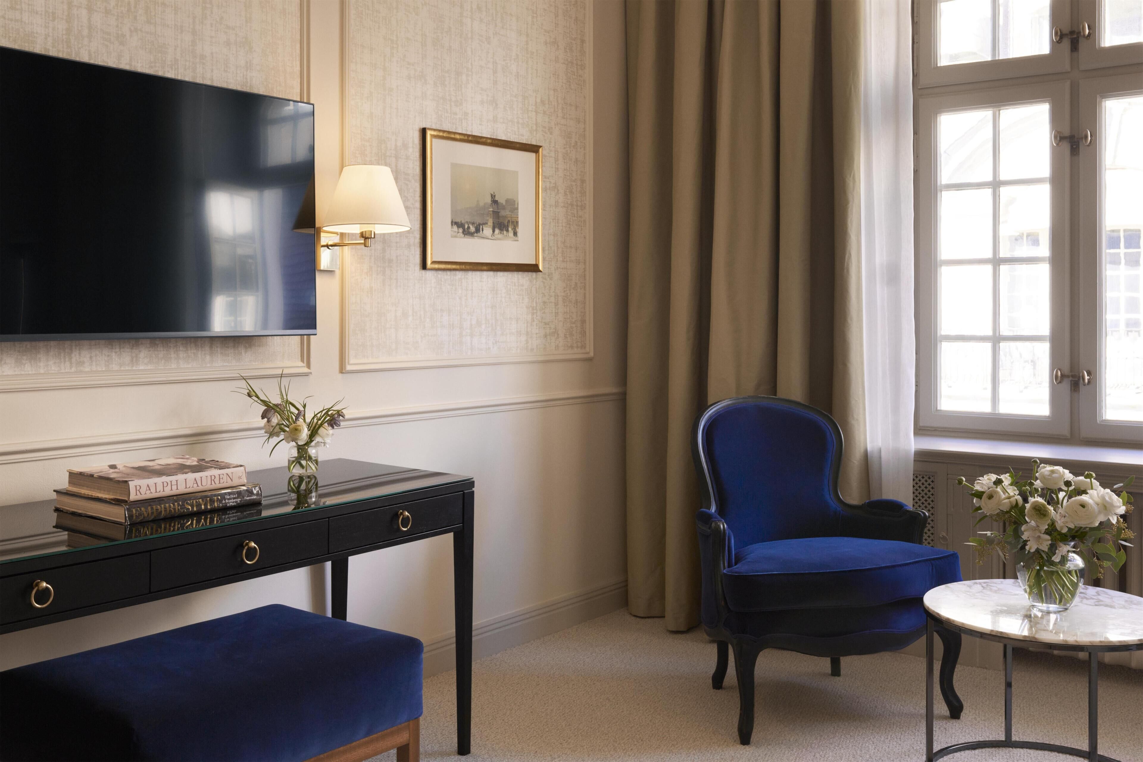 A room with a flat-screen TV, a desk with books, a chair, a small table with flowers, and a window with curtains.