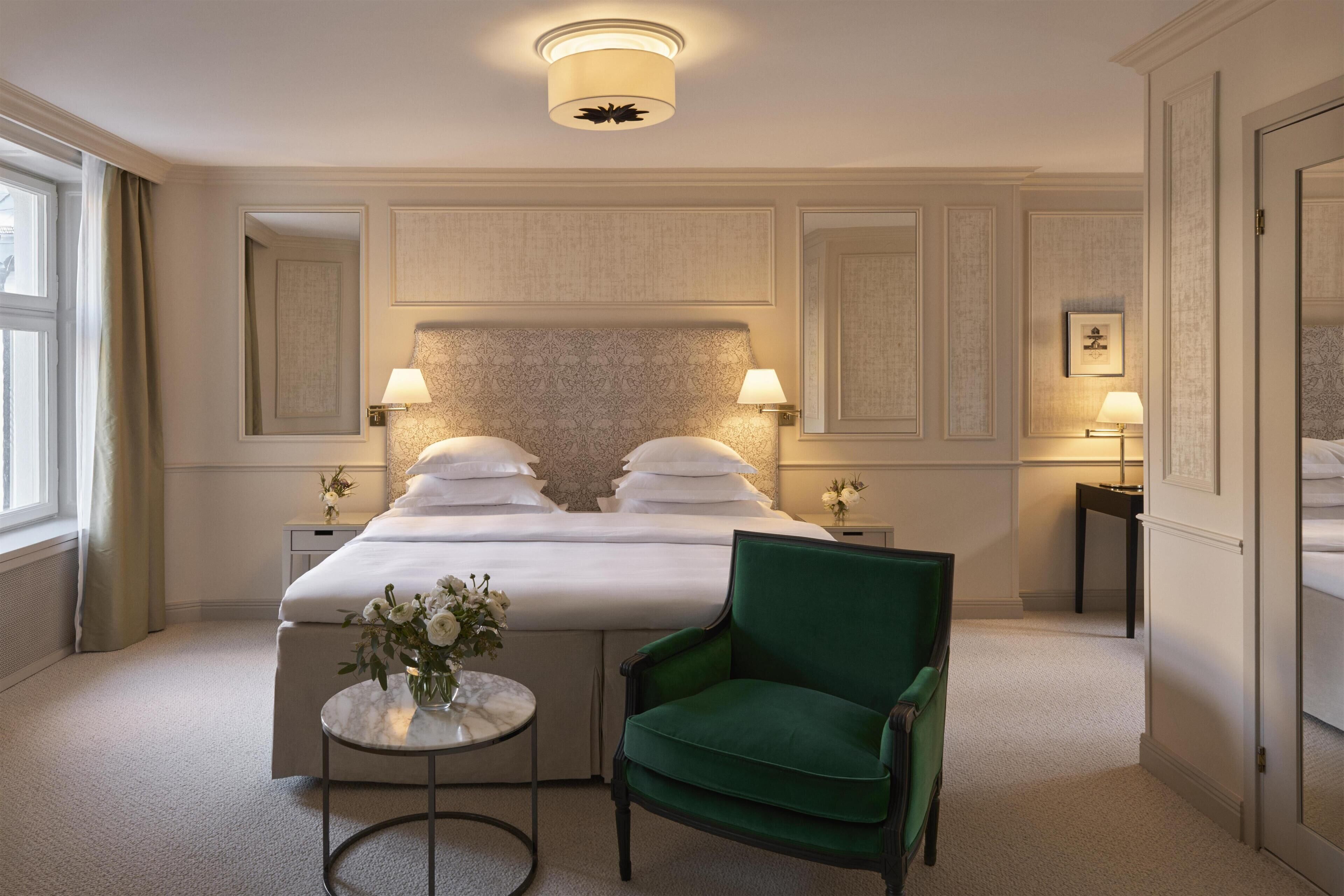 A hotel room with a large bed, two bedside tables with lamps, a green armchair, a small round table with a vase, and a mirror on the wall.