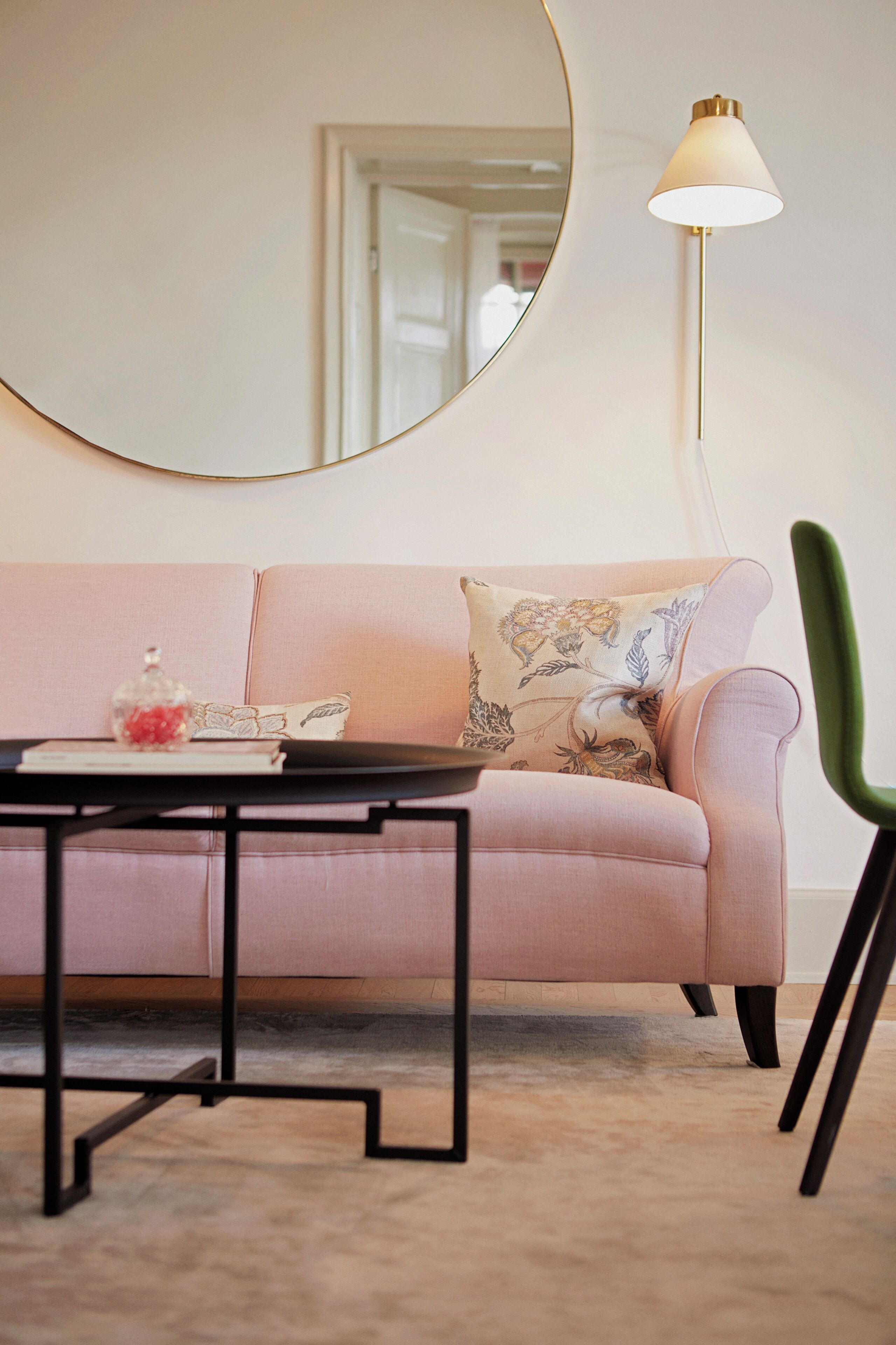 Un sofá rosa con un espejo redondo encima, una mesita con objetos decorativos y una silla verde.