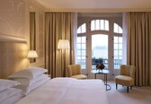 A hotel room with a large bed, two armchairs, a small table with a vase, and a view through a large window.