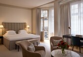A hotel room with a bed, two bedside lamps, a desk with a chair, a small table, and a vase with flowers.