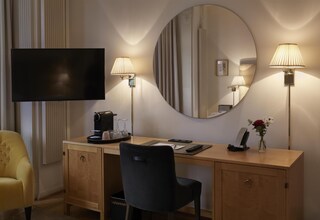 A hotel room with a desk, two lamps, a flat-screen TV, a mirror, a chair, a coffee maker, a vase with flowers, and a cabinet.