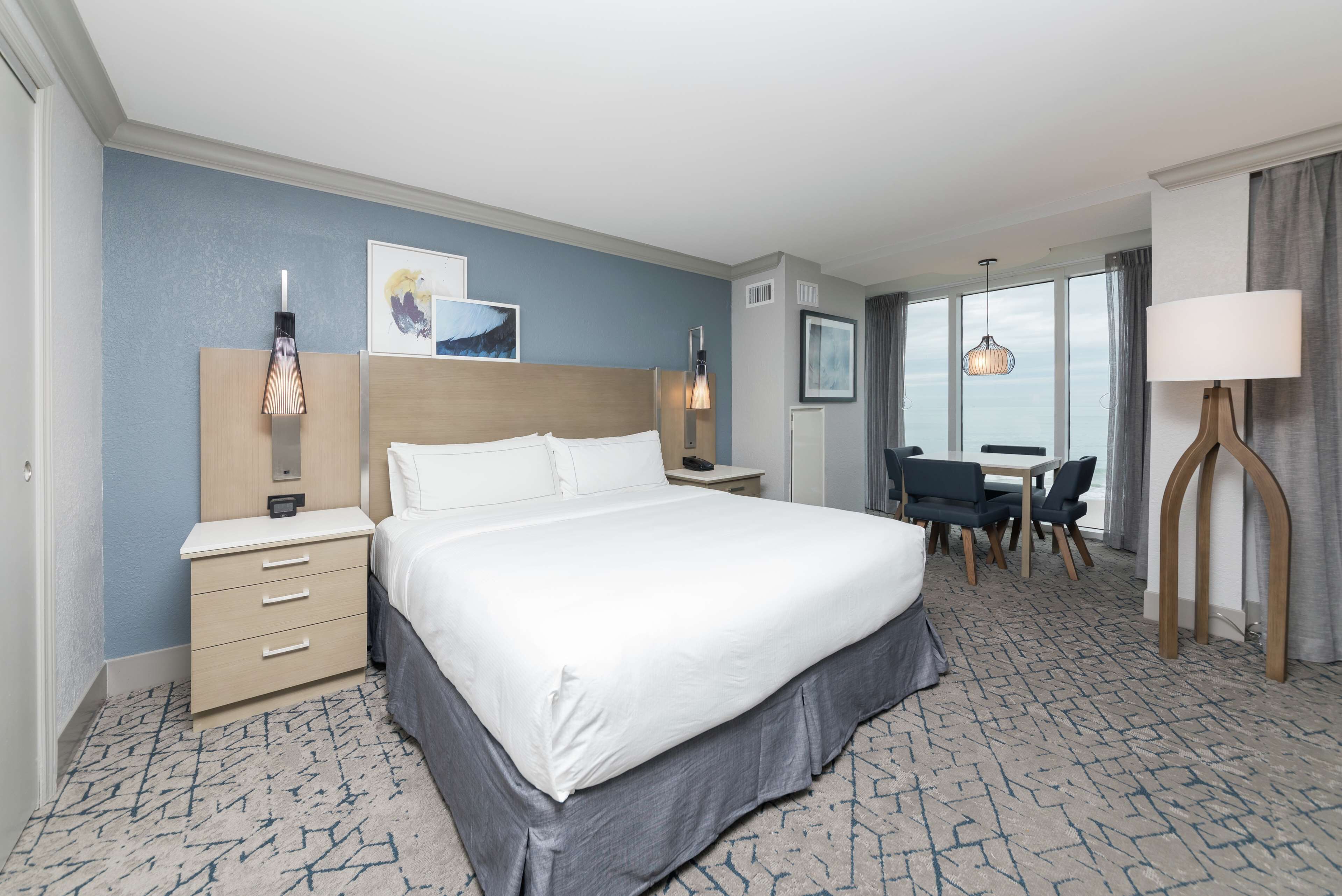 A hotel room with a large bed, bedside tables, a desk with chairs, and a view of the sea through large windows.