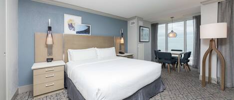A hotel room with a large bed, bedside tables, a desk with chairs, and a view of the sea through large windows.