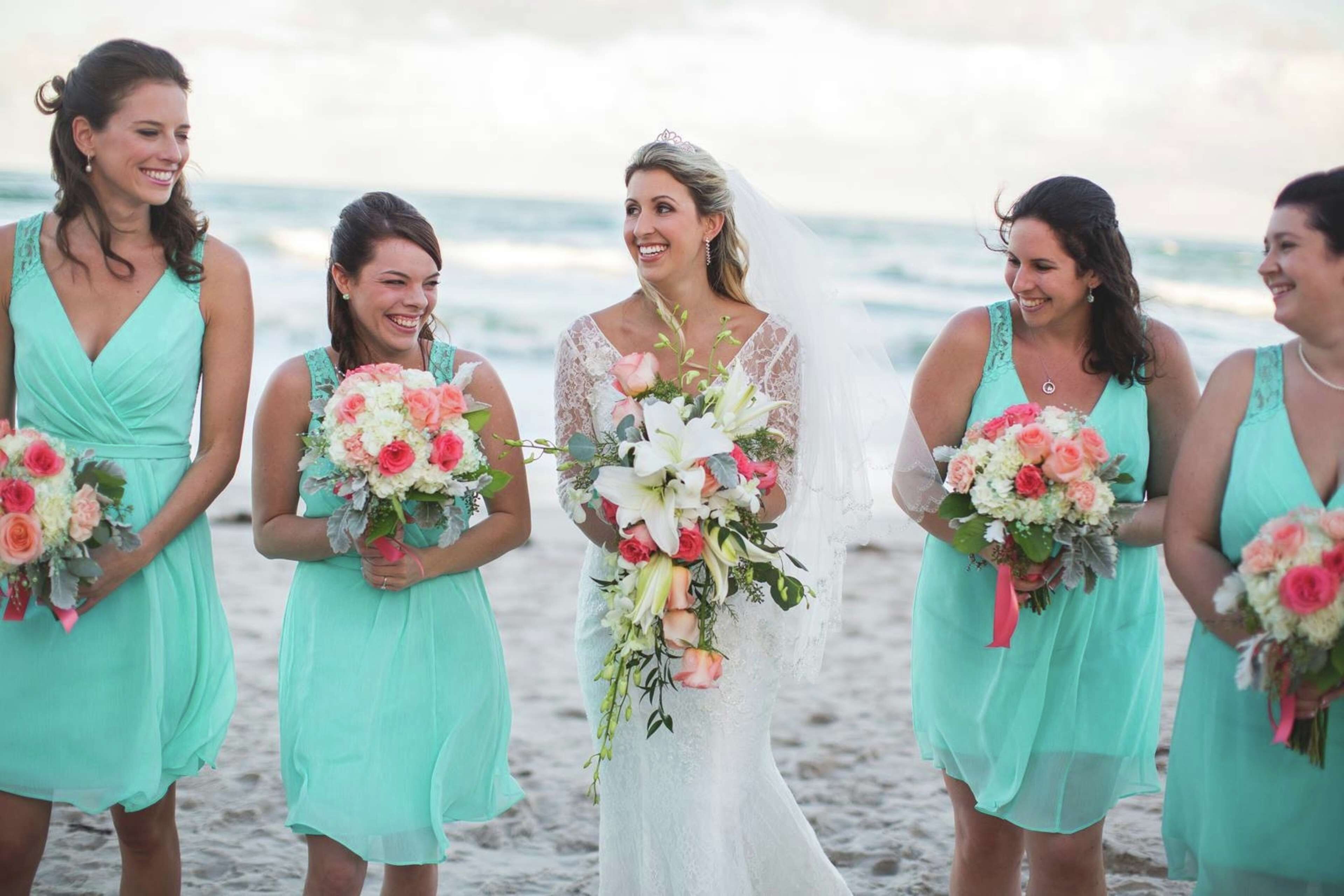 A wedding ceremony on a beach with a bride in a white dress and bridesmaids in matching teal dresses holding bouquets.