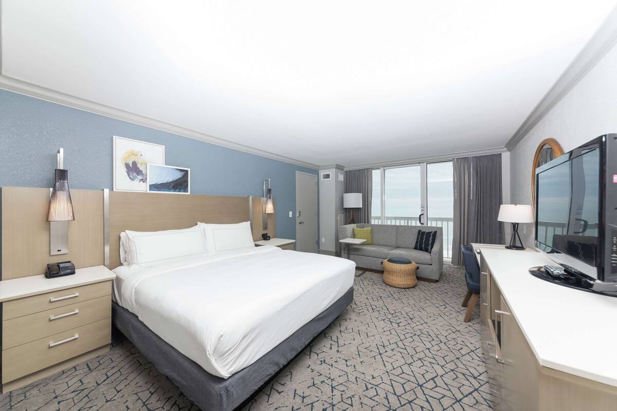 A hotel room with a large bed, a desk with a TV, a sofa, and a window with a view.