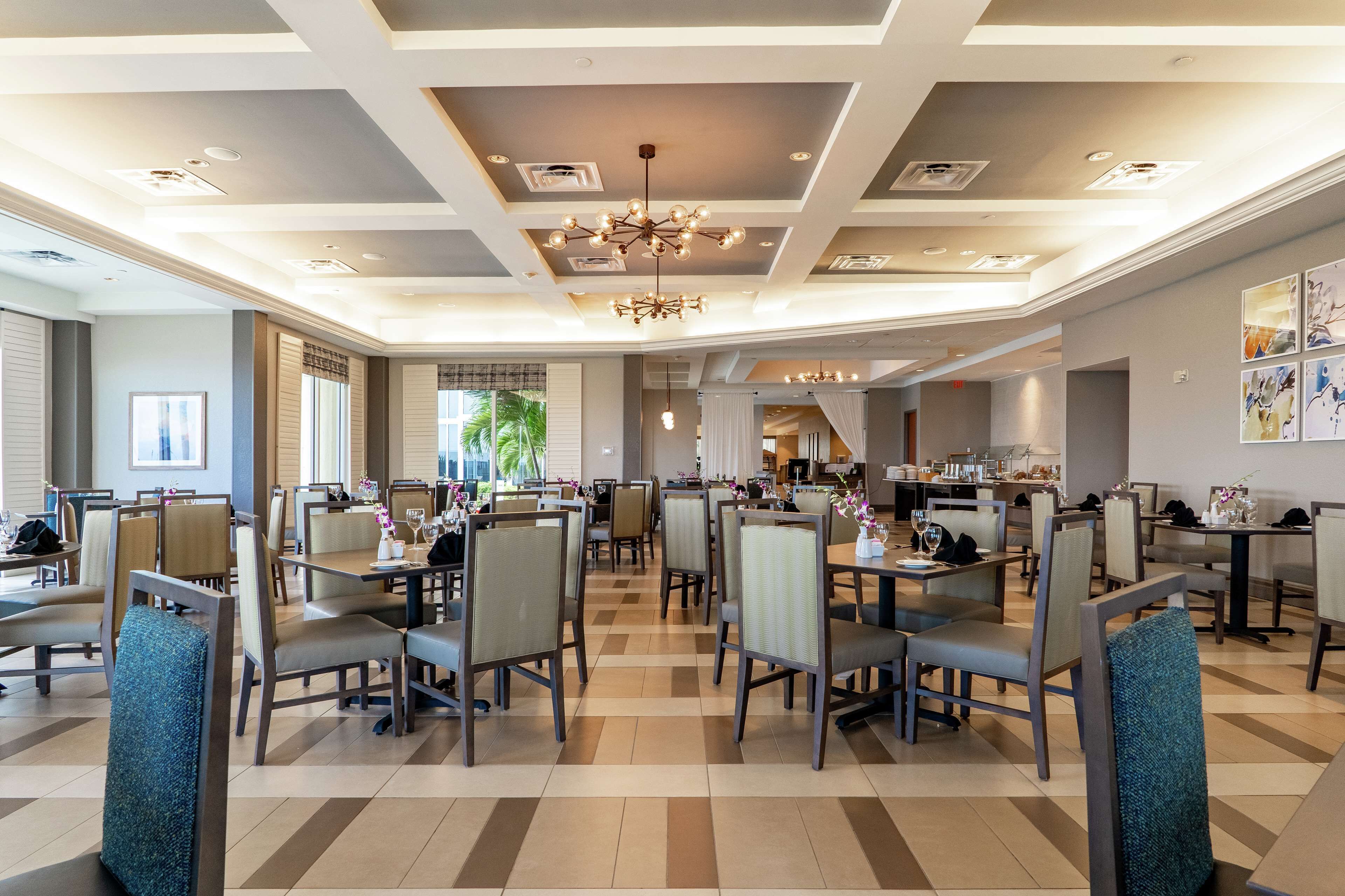 A spacious dining area with multiple tables and chairs, a modern chandelier, and a view of the kitchen in the background.
