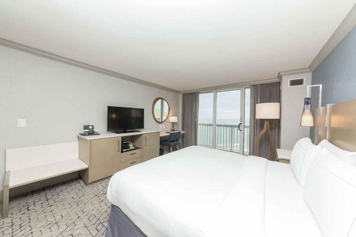 A hotel room with a large bed, a desk with a TV, a chair, a small table, a lamp, and a view of the ocean through the balcony doors.