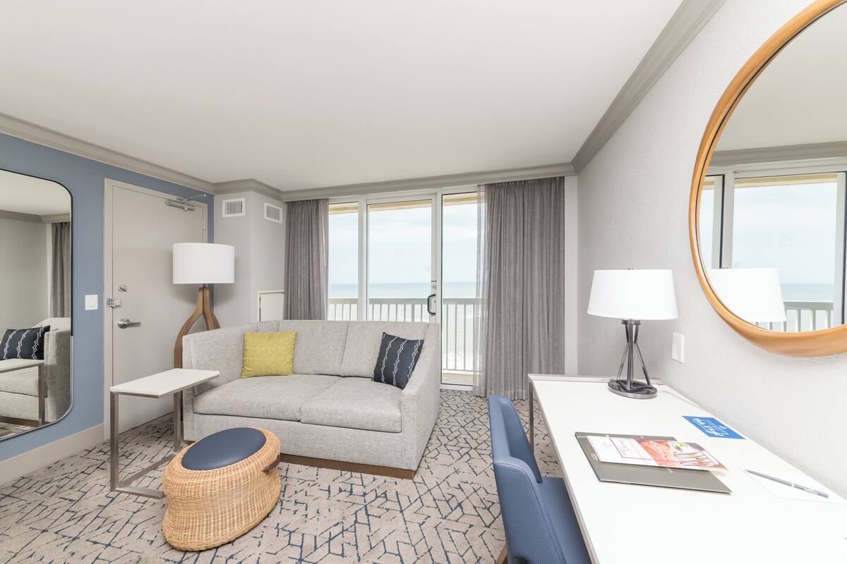 A hotel room with a sofa, a small table, a chair, a wicker ottoman, a desk with a lamp, and a large mirror.