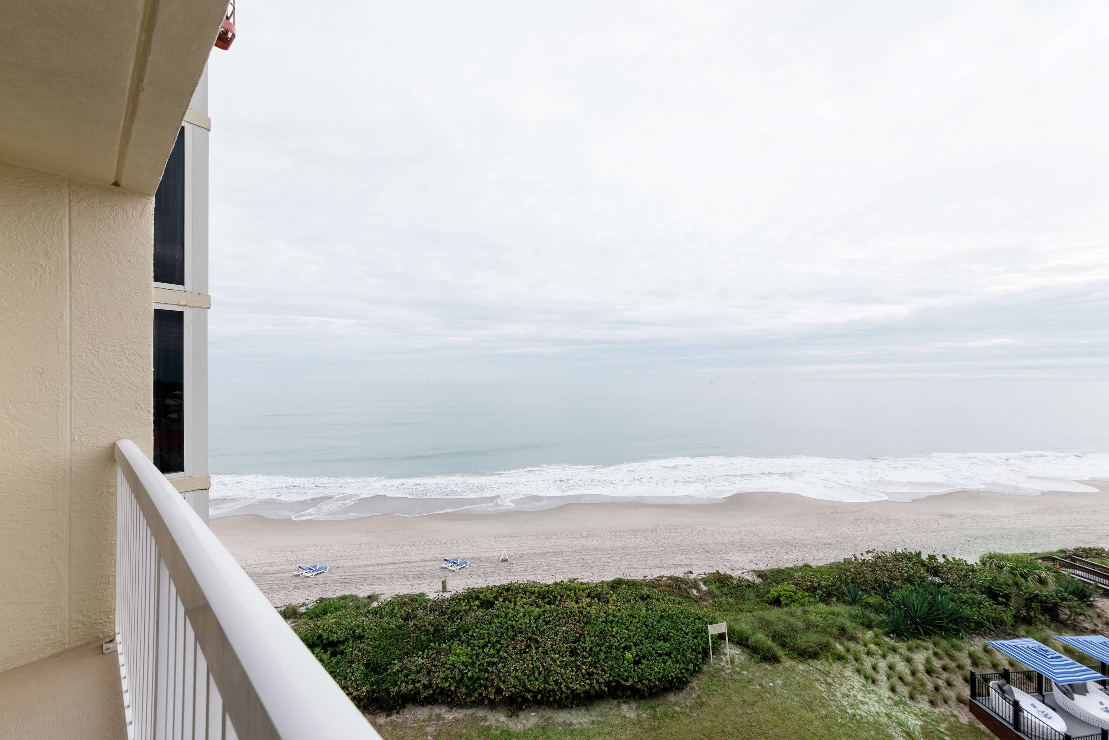 A balcony overlooking a beach with waves, a clear sky, and some greenery.