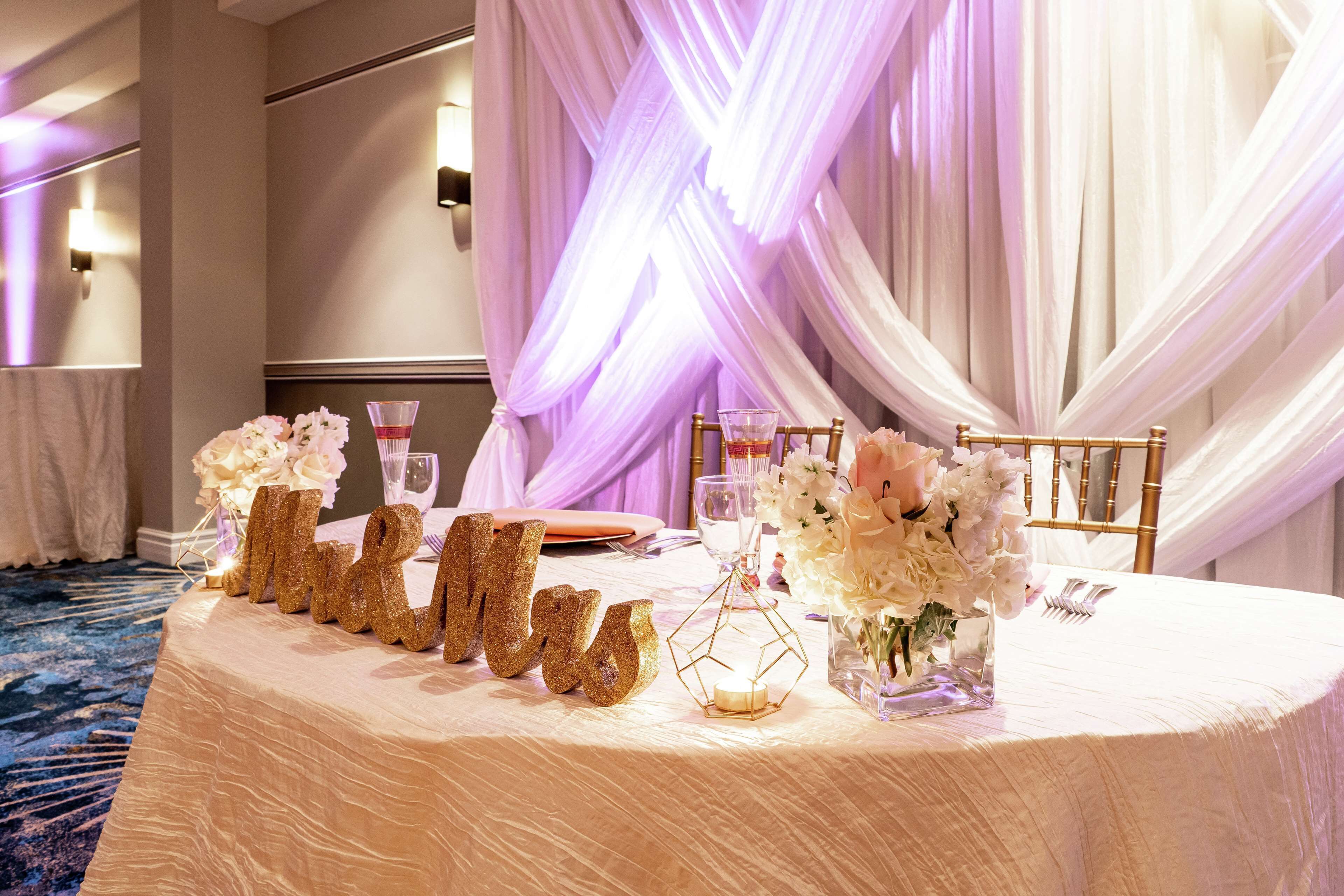 A wedding reception table with a white tablecloth, gold glitter letters spelling "MRS", floral centerpieces, and a gold chair.