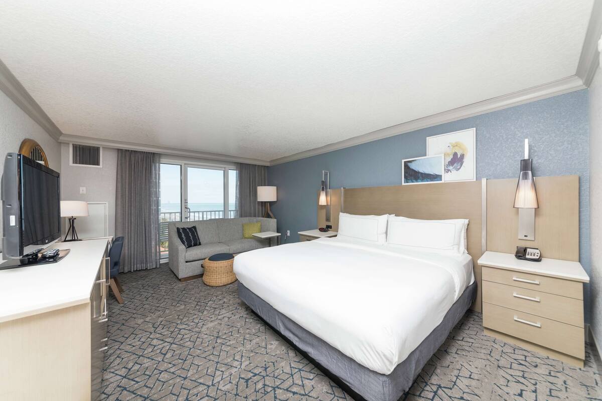 A hotel room with a large bed, a desk, a sofa, a TV, and a balcony with a view.