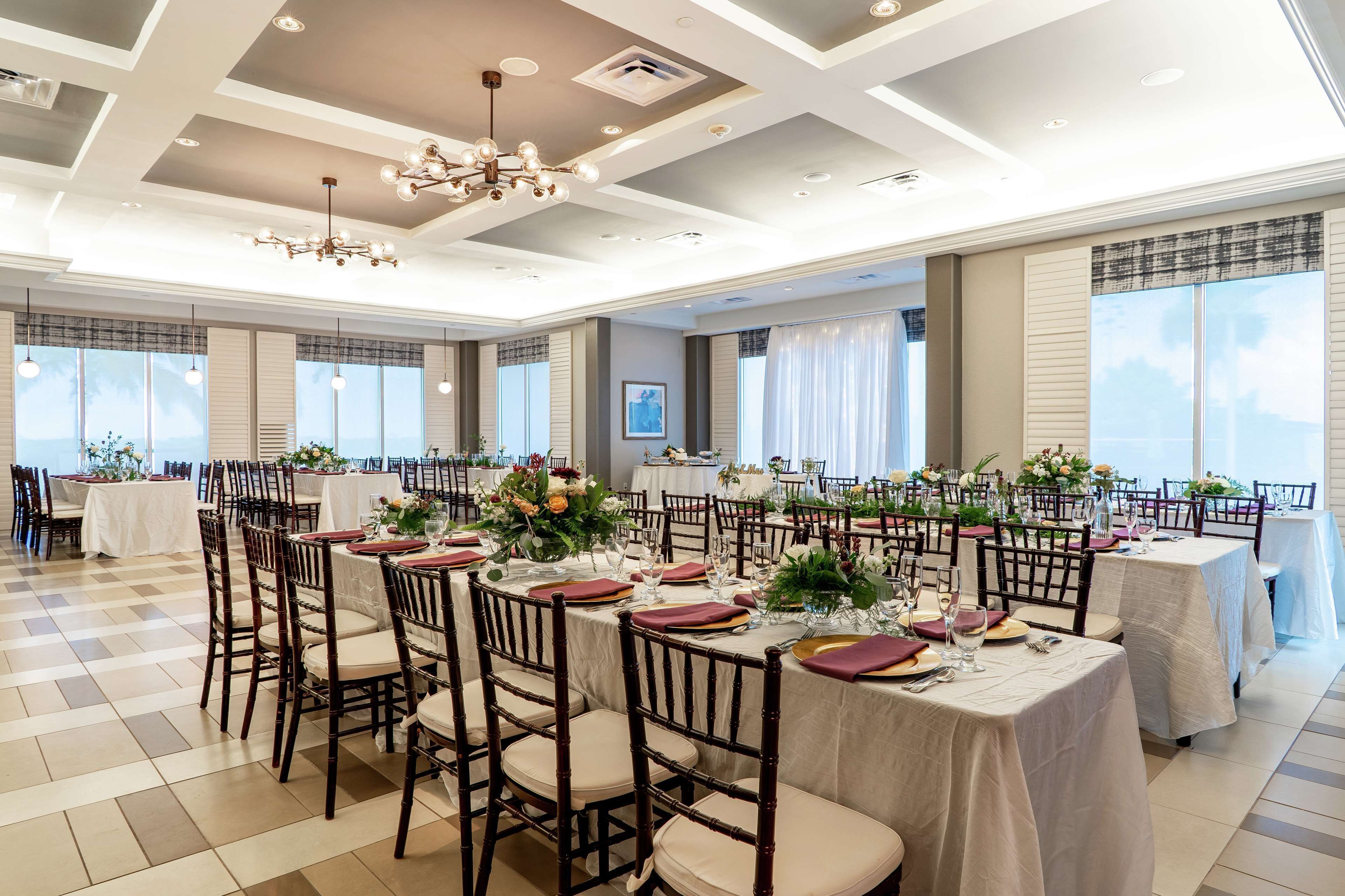 A spacious banquet hall with multiple round tables, each set with white tablecloths, red napkins, and floral centerpieces.