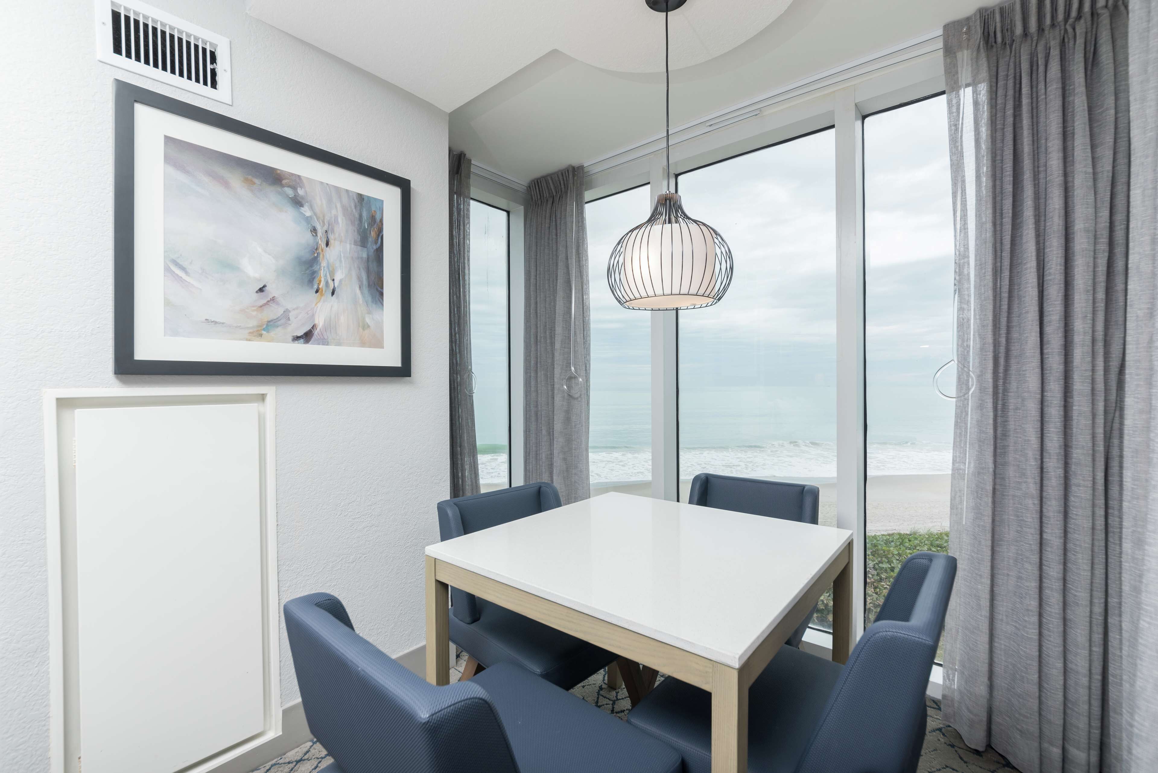 A dining area with a white table and blue chairs, large windows with a view of the ocean, a framed abstract painting, and a hanging pendant light.