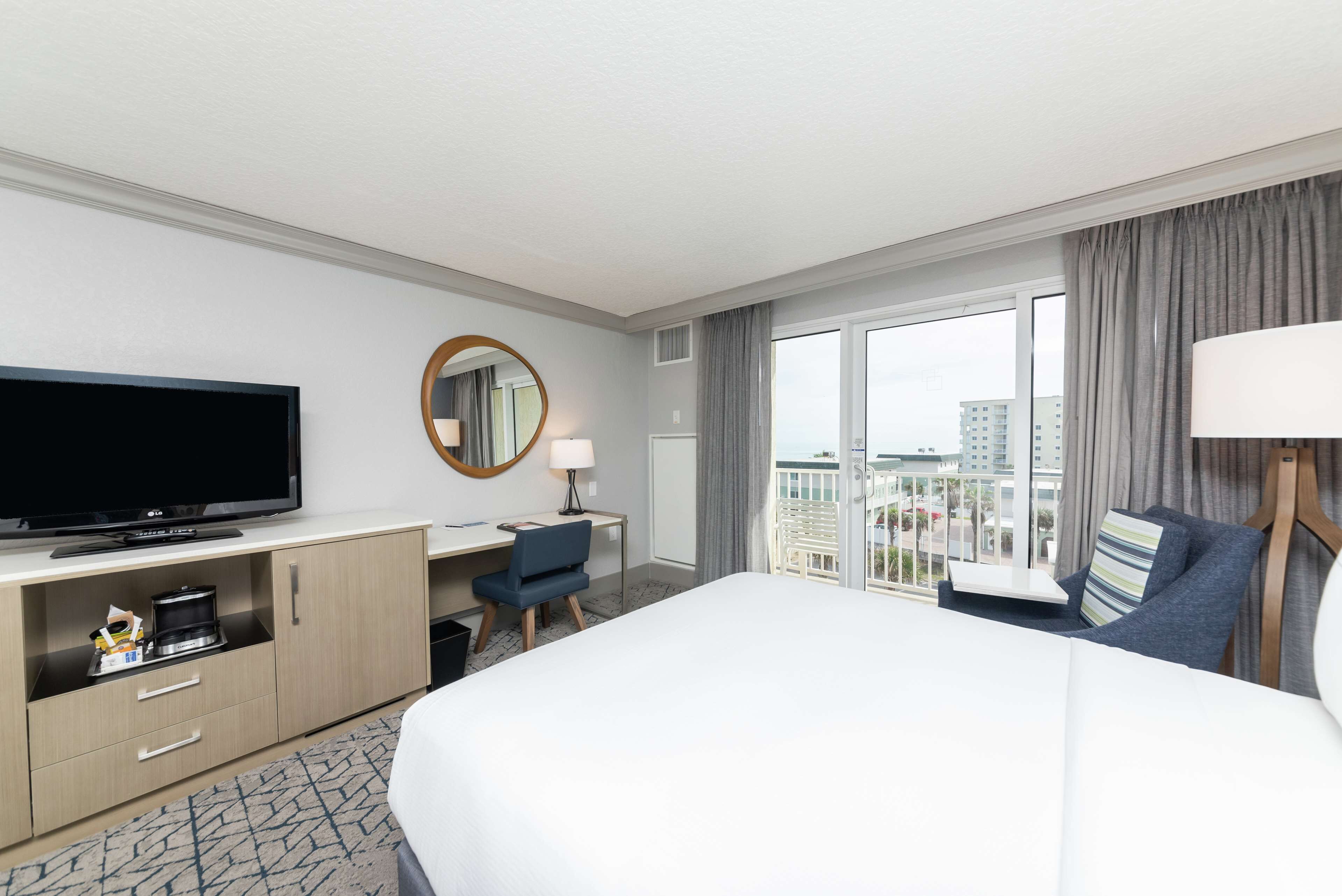 A hotel room with a large bed, a desk with a chair, a television, a mirror, and a view of the city through the window.