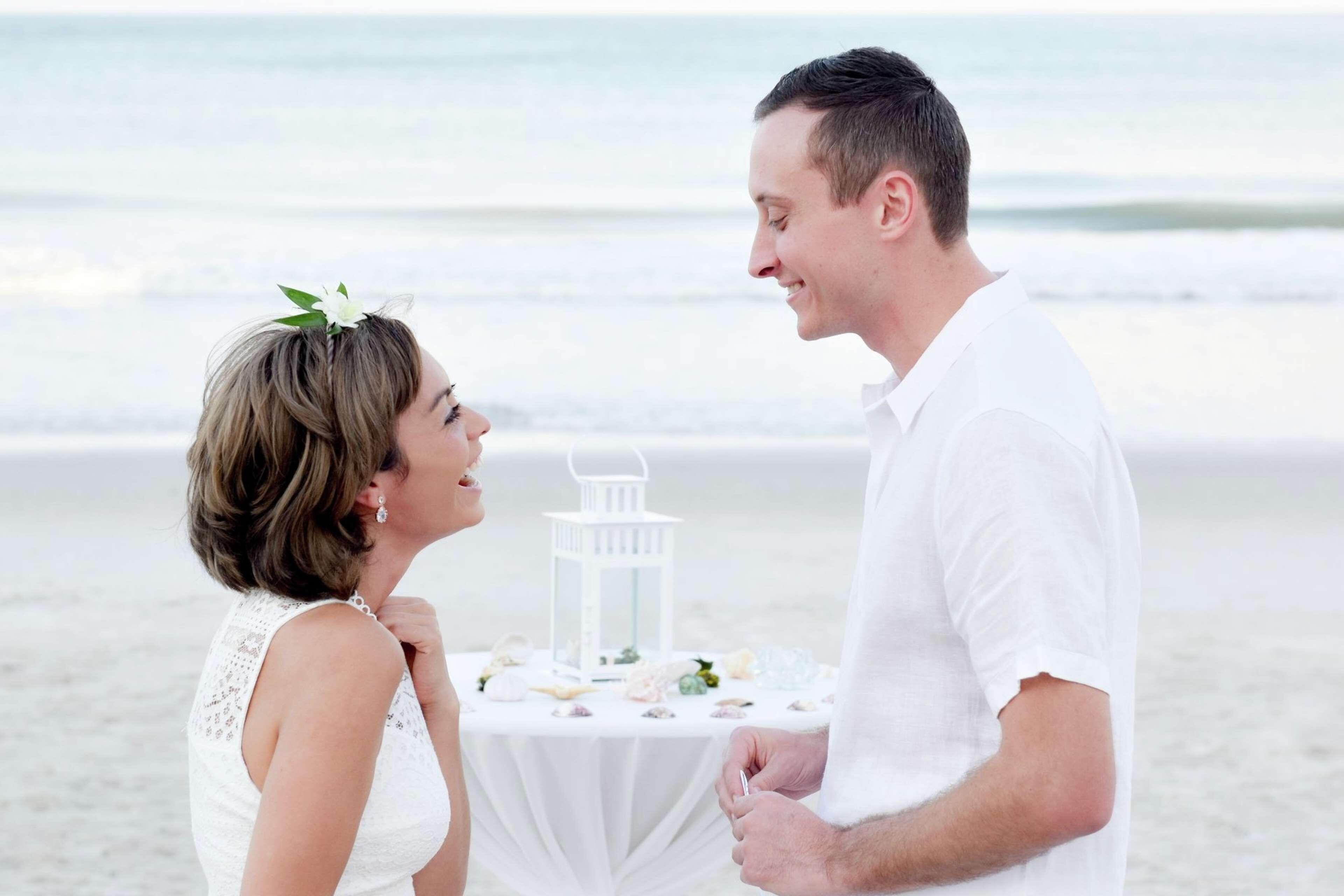 A couple standing at a table on a beach with a white lantern and floral decorations.