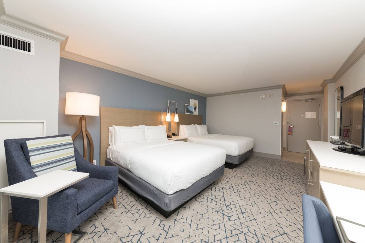 A hotel room with two beds, a desk, a chair, a lamp, and a bathroom door.