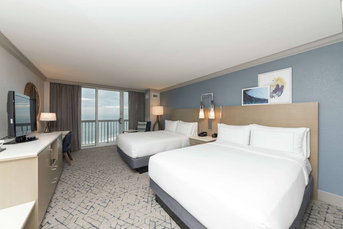 A hotel room with two beds, a desk, a chair, a TV, and a balcony with a view.
