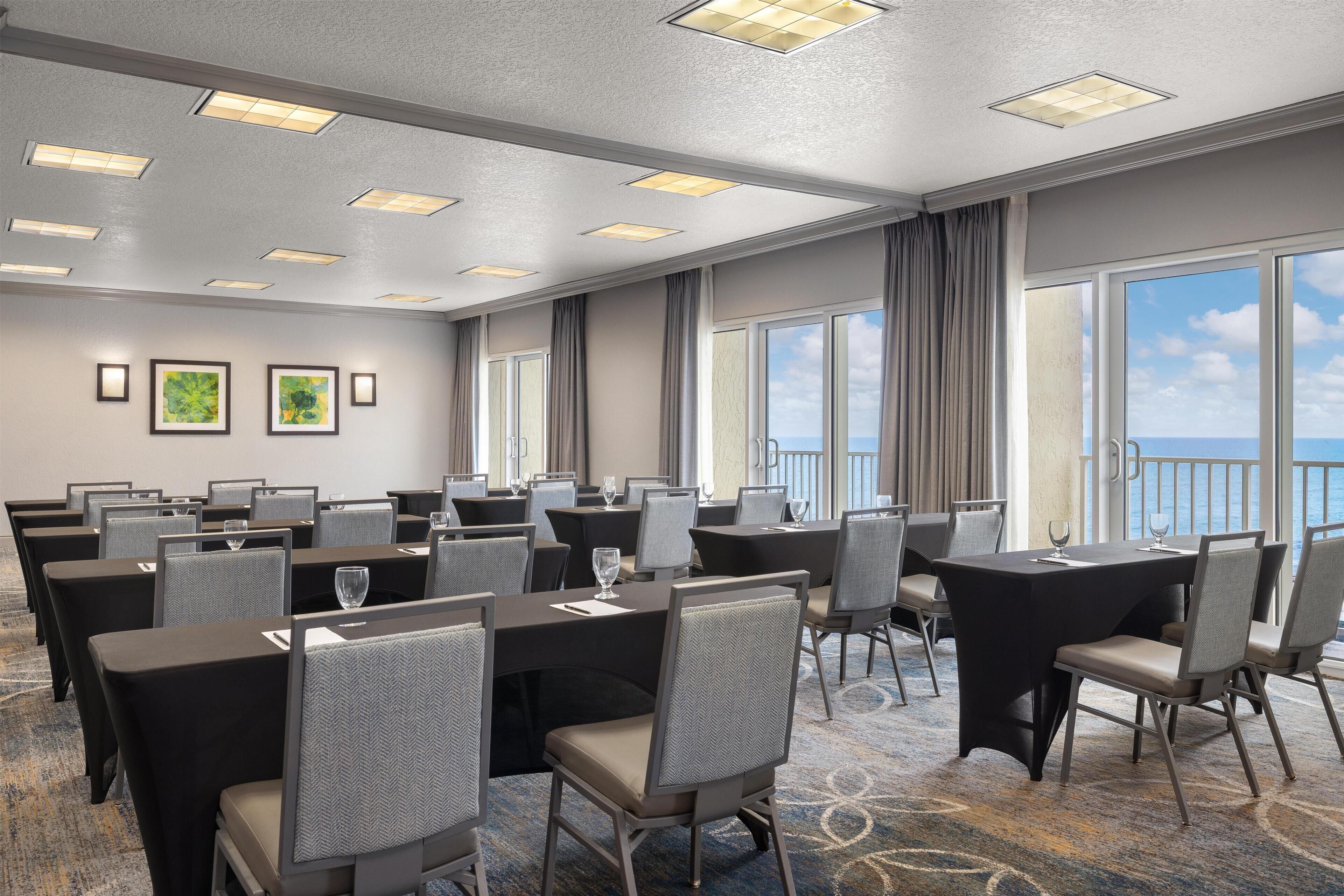 A conference room with multiple tables and chairs, large windows with a view of the sea, and a ceiling with recessed lighting.