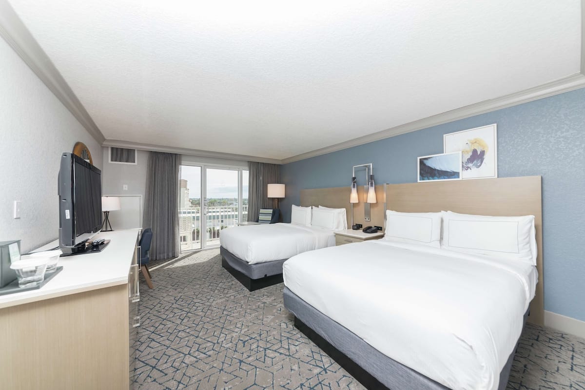 A hotel room with two beds, a desk, a TV, and a large window with a view.
