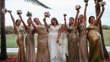 A group of women in formal attire, including a bride, standing outdoors with palm trees and grass in the background.