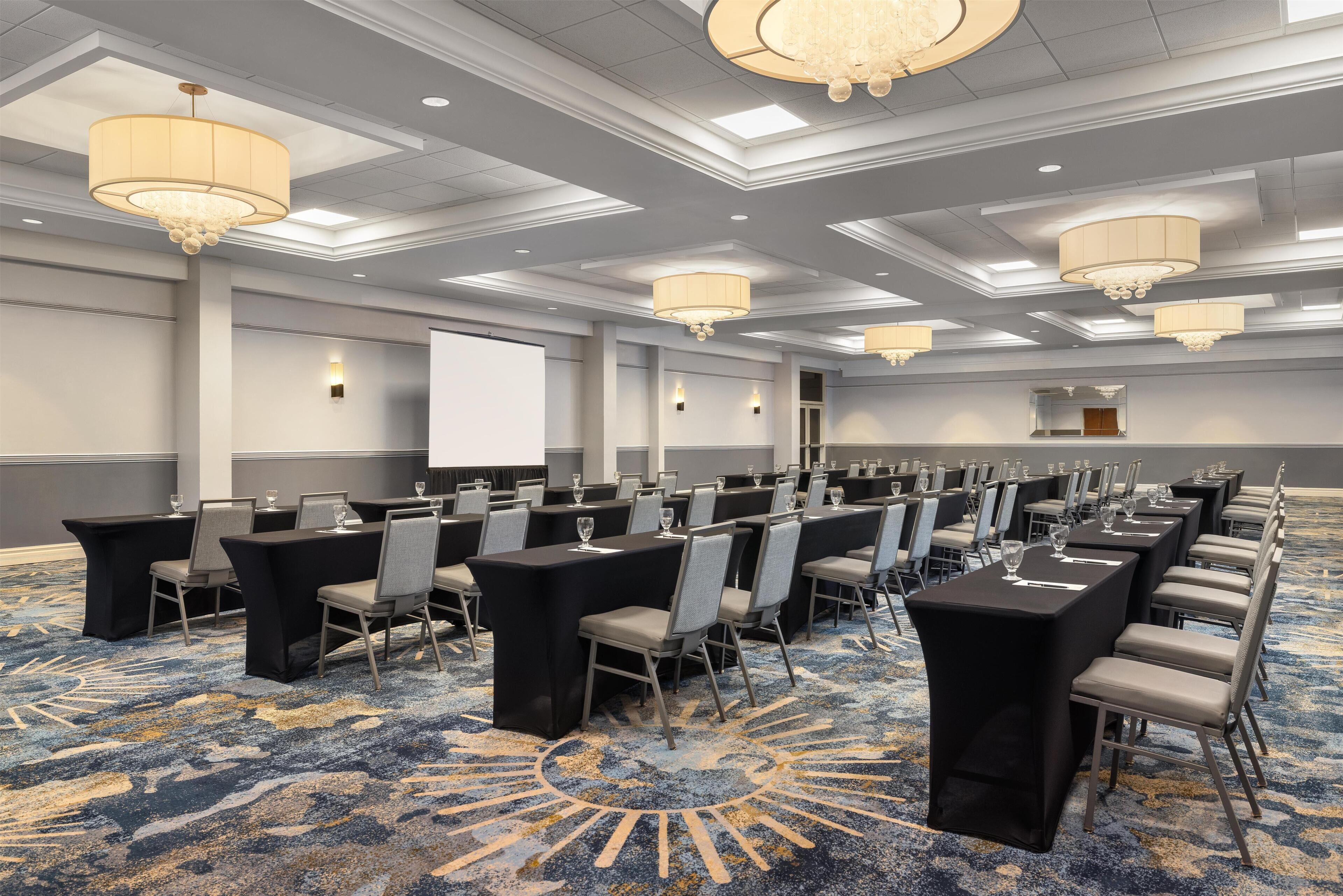 A conference room with multiple tables and chairs, a projector screen, and chandeliers.