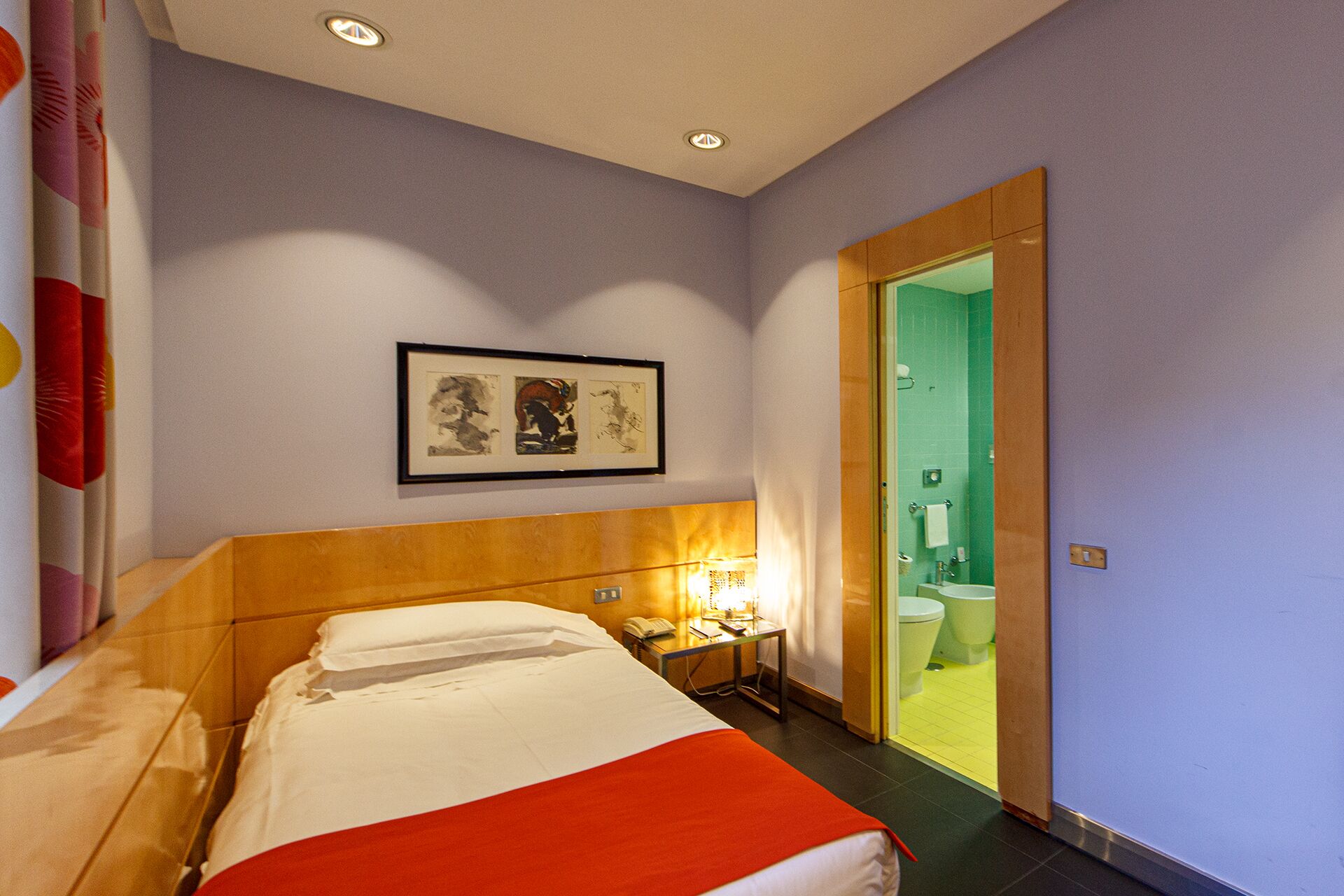 A hotel room with a bed, a nightstand, a framed picture on the wall, and a bathroom visible through an open door.