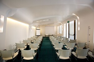 Una sala conferenze con file di sedie bianche, uno schermo per proiezioni e un tavolo con una bottiglia d'acqua.