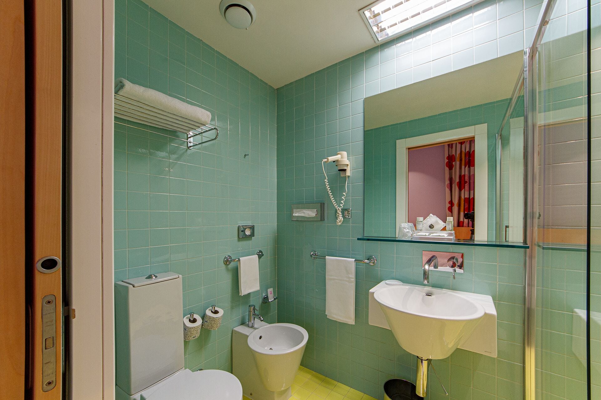 A bathroom with green tiles, a white toilet, a bidet, a sink, and a mirror with a glass enclosure.