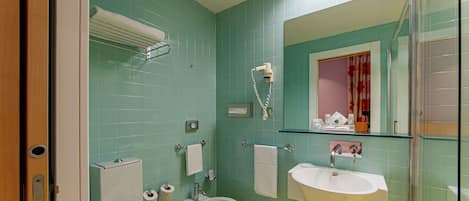 Un baño con azulejos verdes, un inodoro blanco, un bidé, un lavabo y un espejo con cerramiento de vidrio.