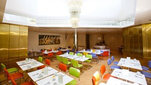 A spacious dining area with multiple tables and chairs, a chandelier, and a high ceiling with natural light.
