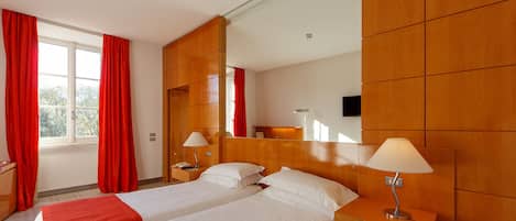 Habitación de hotel con dos camas, un espejo grande, paneles de madera, un escritorio con lámpara y una ventana con cortinas rojas.