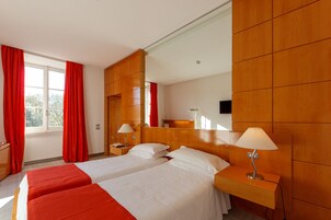 Una camera d'albergo con due letti, un grande specchio, pannelli in legno, una scrivania con lampada e una finestra con tende rosse.