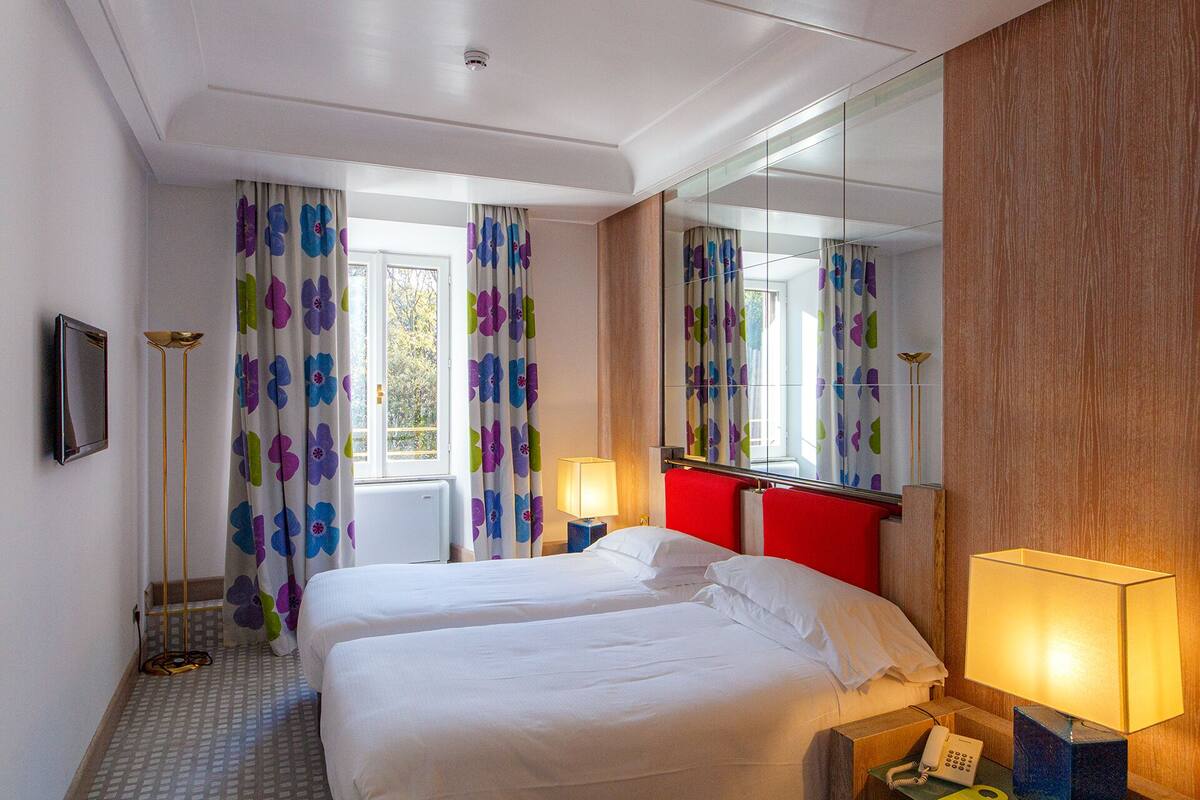 A hotel room with two beds, a large mirror, a red accent wall, and a window with curtains.