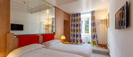 Una habitación de hotel con dos camas, un espejo grande, un televisor y una ventana con cortinas.
